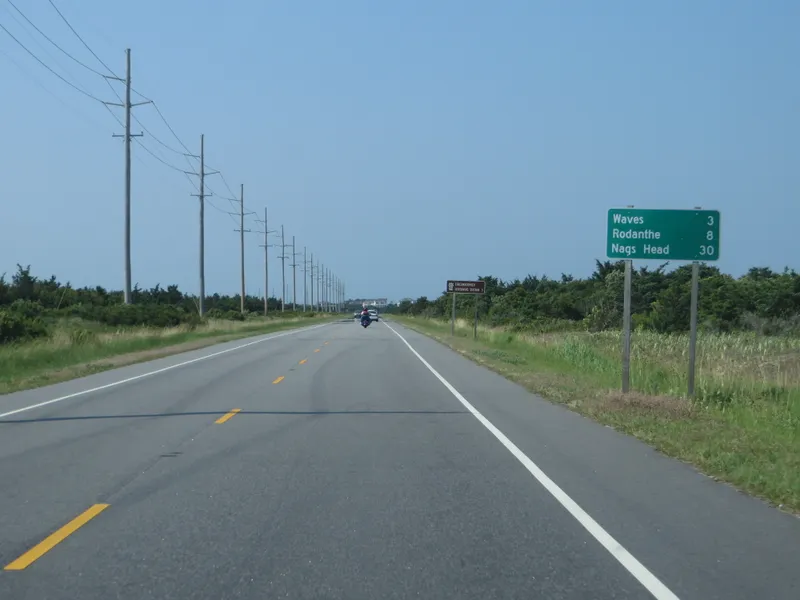 Highway 12 road through Cape Hatteras National Seashore with a sign showing 3 miles to the village of Waves, beach and dunes visible