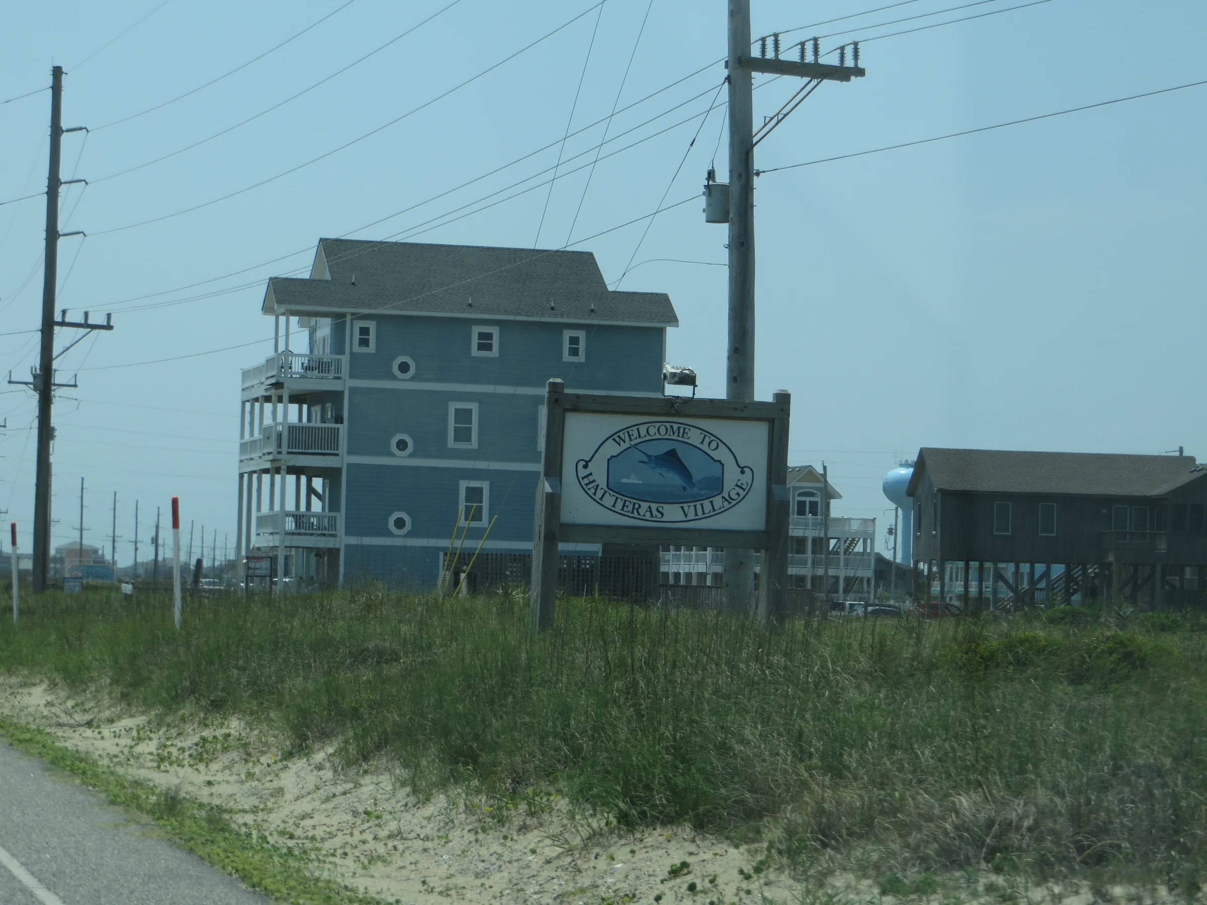 Welcome to Hatteras Village sign along Highway 12 on the Outer Banks with beach houses and maritime vegetation